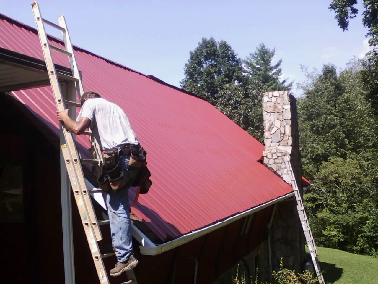 Roofer on a ladder beside a red metal roof and stone chimney — Soffit & Fascia Repair by Roofing Brothers in Sandy Hook
